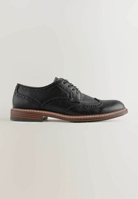 Chaussure habillée brogue en cuir noir avec perforations wingtip, talon empilé en bois et fermeture lacée sur fond gris clair.