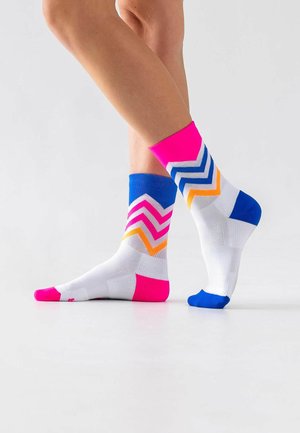 Beine mit unpassenden weißen Sportsocken mit farbigen Zickzackstreifen in Pink, Blau, Orange und Flächen in kräftigem Pink und Blau.