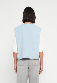 Μπλε γιλέκο από φλις με υφή, με λευκά πλεκτά μανίκια και χαλαρή εφαρμογή. Συνδυασμένο με γκρι παντελόνια. Απλός, μοντέρνος σχεδιασμός.