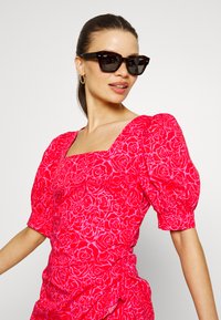 Vestido floral vermelho com mangas curtas bufantes e decote quadrado; apresenta um padrão de rosas em tons de rosa mais claros. Usado com óculos de sol escuros.