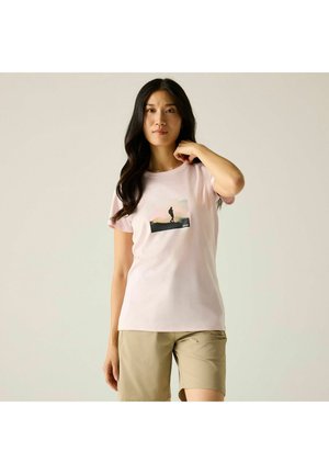 Femme aux longs cheveux foncés portant un t-shirt rose clair avec un motif silhouette et un short beige, debout avec une main près du cou.