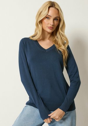 Vrouw met lang blond haar die een donkerblauwe V-hals trui en lichtblauwe jeans draagt, staand tegen een effen lichte achtergrond.