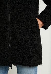 Cappotto in pile nero con tessuto texturizzato e riccio, dotato di cerniera anteriore, tasche laterali e orlo dritto.