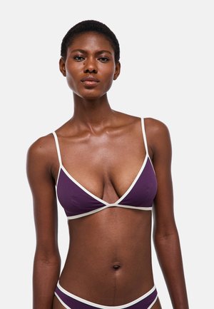 Femme noire portant un bikini violet avec bordure blanche, debout devant un fond gris clair uni.