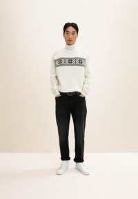 Homme debout, les mains dans les poches, portant un pull à col roulé blanc à motifs, un jean noir et des baskets blanches, sur un fond beige uni.