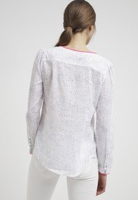 Weißes Langarmhemd mit lila Punktmuster, runder Saum und pinken Akzenten am Kragen und an den Manschetten. Glatte Stoffoberfläche.