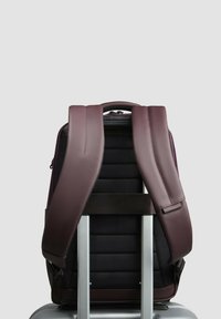 Zaino con spallacci larghi viola, corpo nero con trama, pannello posteriore imbottito e maniglia superiore, montato su un carrello per bagagli argentato.