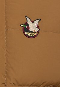Brun pufferjakke-sektion med et broderet patch af en flyvende and og teksten "CHEVIGNON TOG'S" i en cirkulær sort og rød kant.