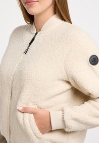 Beige, fluffy fleece jacket with a zip front, ribbed cuffs, side pockets, and a black logo patch on the sleeve. Soft texture, loose fitting.