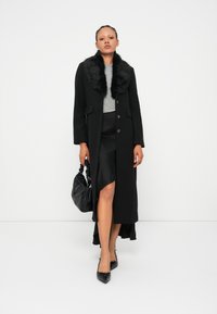 Cappotto di lana nera con colletto in pelliccia sintetica, vestibilità sagomata, bottoni frontali e tasche laterali. La modella tiene in mano una borsa nera con texture.