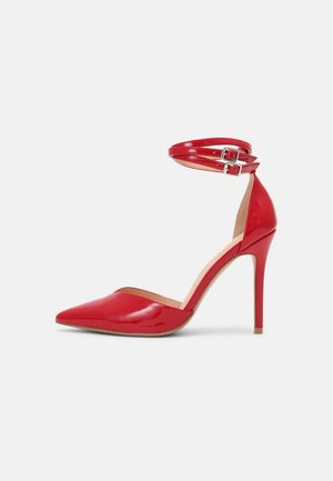 Chaussure à talon haut en cuir verni rouge avec bout pointu et double bride à la cheville avec boucles argentées sur fond blanc.