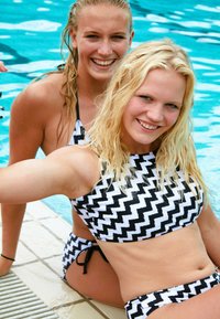 Two women are sitting by a pool in matching black and white zigzag patterned swimsuits. The fabric is smooth with wet hair visible.