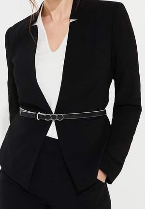 Kvinde iført sort, skræddersyet blazer over hvid V-hals top, med slank sort bælte med tre sølvfarvede ringe ved taljen.