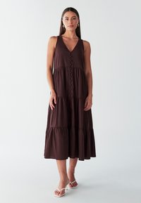 Brun maxikjole med lag på lag-effekt, V-udskæring og knapper i midten; lavet af blødt stof; har tynde stropper og en flydende silhuet.