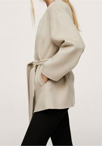 Beige wikkeljas met een ceintuur, lange mouwen, zijzakken en een zachte textuur, uitgevoerd in een eenvoudig, recht gesneden ontwerp.