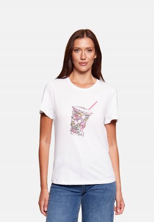 Donna che indossa una T-shirt bianca a maniche corte con design di bicchiere da bevanda decorato in rosa e argento, abbinata a jeans blu, in piedi davanti a uno sfondo semplice.