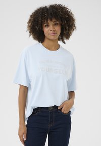 T-shirt bleu clair à manches courtes avec un col rond. Présente un graphique textuel en lettres métalliques qui dit "LE VRAI CROISSANCE EST D'ÊTRE PLUS SOI-MÊME."