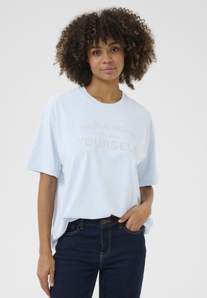 T-shirt bleu clair à manches courtes avec un col rond. Présente un graphique textuel en lettres métalliques qui dit "LE VRAI CROISSANCE EST D'ÊTRE PLUS SOI-MÊME."