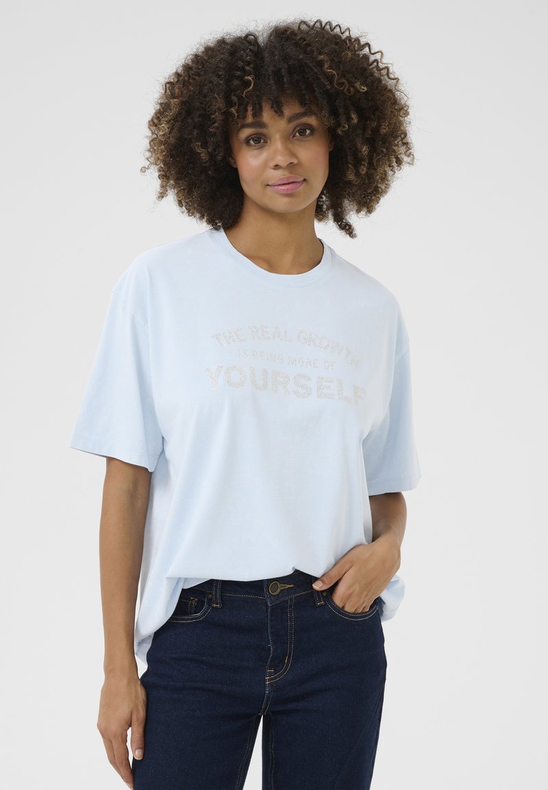 Ljusblå t-shirt med kort ärm och rund halsringning. Har en textgrafik med metallisk skrift som lyder "DEN VERKLIGA TILLVÄXTEN ÄR ATT VARA MER AV DIG SJÄLV."