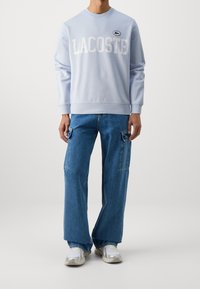 Felpa azzurra chiara con la scritta "LACOSTE" in lettere bianche. Abbinata a jeans blu larghi con tasche laterali e sneakers grigie.