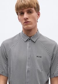 Cămașă cu mâneci scurte și guler, în dungi verticale alb-negru, cu un logo subtil pe pieptul stâng. Textură de țesătură netedă.