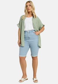 Ljusgrön, kortärmad skjorta med knappar över en vit t-shirt, i kombination med ljusblå denimbermuda-shorts och vita sandaler.