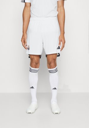 Mężczyzna sportowiec ubrany w białe spodenki piłkarskie Realu Madryt, długie białe skarpetki Adidas z czarnymi paskami oraz białe korki, stojący z rozluźnionymi ramionami.