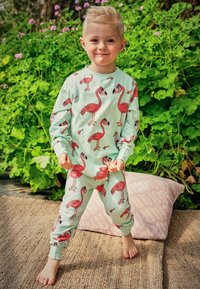 Pajamas made of soft mint fabric featuring red flamingo prints, long sleeves, cuffs, and matching pants. Barefoot on a textured rug.
