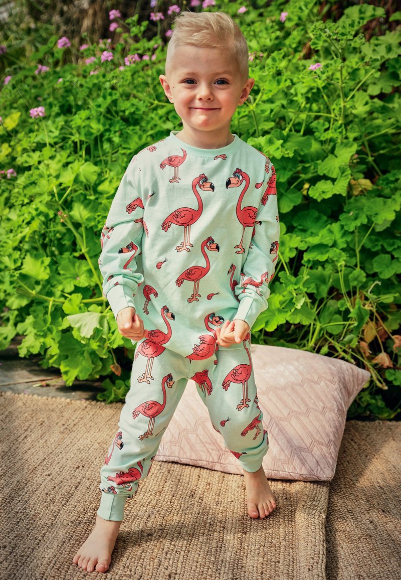 Pajamas made of soft mint fabric featuring red flamingo prints, long sleeves, cuffs, and matching pants. Barefoot on a textured rug.
