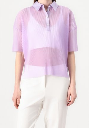Femme portant un polo manches courtes en lavande transparente par-dessus un haut blanc, associé à un pantalon blanc taille haute sur un fond uni.