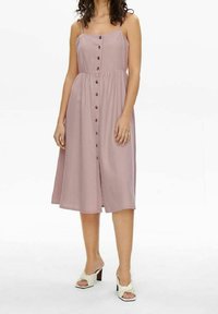 Ljusrosa, klänning i midi-längd med rak halsringning, tunna axelremmar och knappstängning fram; tyget verkar slätt och lätt.