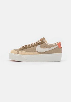 Niskie beżowo-brązowe buty sportowe z białym logo Nike Swoosh, beżowymi zamszowymi akcentami, białą podeszwą na platformie i pomarańczową zakładką na pięcie.