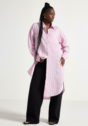 Donna con trecce lunghe che indossa una camicia oversize a righe rosa con bottoni e pantaloni neri a gamba larga, in posa su uno sfondo bianco semplice.