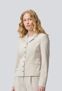 Beige skräddarsydd blazer med långa ärmar, notched lapeller och guldknappar. Har två framfickor och en skarp, strukturerad design.