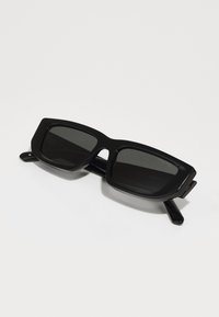 Ochelari de soare negri, supradimensionați, cu un cadru din plastic gros, lentile închise la culoare și un design unghiular. Prezintă brațe drepte și o marcă subtilă.