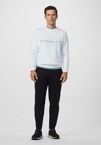 Uomo in piedi che indossa una felpa leggera blu con il testo "HACKETT", pantaloni neri e sneakers nere su uno sfondo bianco semplice.