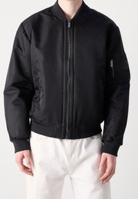 Veste bomber noire avec une finition lisse et brillante, dotée d'une fermeture éclair frontale, d'un col et de poignets côtelés, ainsi que d'une poche zippée sur la manche gauche.