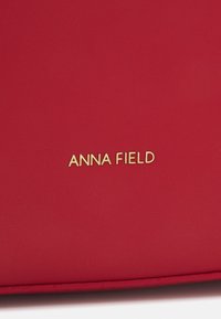 Sac à main synthétique rouge présentant une texture lisse et un logo doré "ANNA FIELD". Design simple avec des bords plats et sans autres ornements.