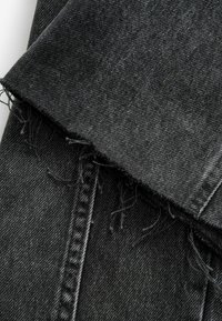 Tissu en denim noir avec un bord effiloché et une couture visible le long de la couture sur un fond blanc.