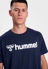 Indigo mėlynas medvilninis marškinėlis su dideliu baltu "hummel" logotipu priekyje, tradiciniu apvaliu kaklu ir trumpomis rankovėmis.