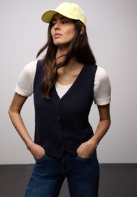 Femme aux cheveux longs bruns portant une casquette jaune, un haut blanc à manches courtes, un gilet noir boutonné et un jean bleu, les mains dans les poches, regardant sur le côté.