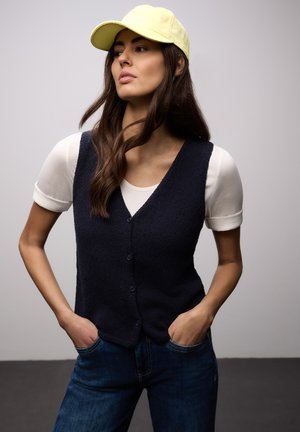 Donna con capelli castani lunghi indossa un berretto giallo, una maglietta bianca a maniche corte, un gilet nero con bottoni e jeans blu, con le mani in tasca, guarda di lato.