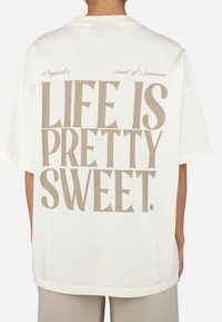 White oversized T-shirt made of cotton, featuring bold beige text on the back: "LIFE IS PRETTY SWEET." Brand logos above the text.