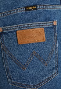 Sininen denim-kangas, jossa on takatasku, jossa on ruskea nahkainen etiketti, jossa on kohokuvioitu "Wrangler". Kaksoisommeltu luo sahalaitakuviota.