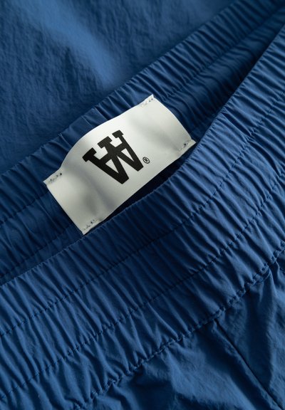 Blaue Hose mit elastischem Taillenbund, bestehend aus einem strukturierten Stoff und einem weißen Etikett mit schwarzem Logo. Der Stoff scheint leicht und glatt zu sein.