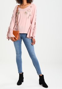 Top lungo a maniche con ricami floreali di un delicato rosa, maniche a campana, abbinato a jeans skinny blu e stivali neri alla caviglia. Borsa a tracolla marrone.