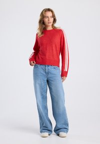 Pull rouge avec des rayures blanches sur les manches, ourlet côtelé, associé à un jean large bleu clair et des chaussures claires, design minimaliste.