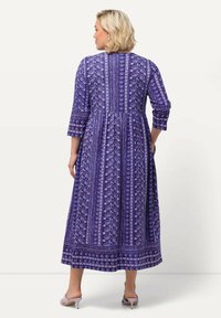 Violetti maksimekko, jossa on kolmennesarleiset hihat, kukkakuosiset ja geometriset kuviot, vyötäröltä tiukka malli ja lentävä helma. Kankaassa on tekstuuria.