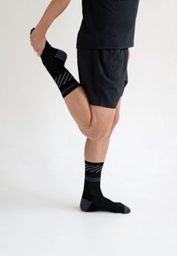 Schwarze Sportsocken mit grauen Akzenten und diagonalen Streifen, getragen von einer Person, die sich dehnt. Der Stoff sieht glatt und elastisch aus.