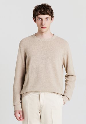 Junger Mann mit kurzen, lockigen braunen Haaren, der einen beigen Strickpullover und crèmefarbene Hosen trägt und mit einer Hand in der Tasche vor einem weißen Hintergrund steht.